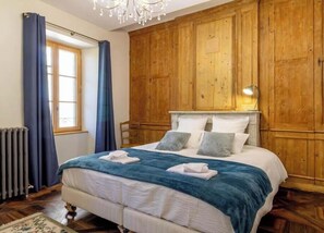 A bedroom with a wooden headboard, a large bed with white bedding, and a window with blue curtains. - Stendhal House - La Chartreuse (LES ECHELLES)