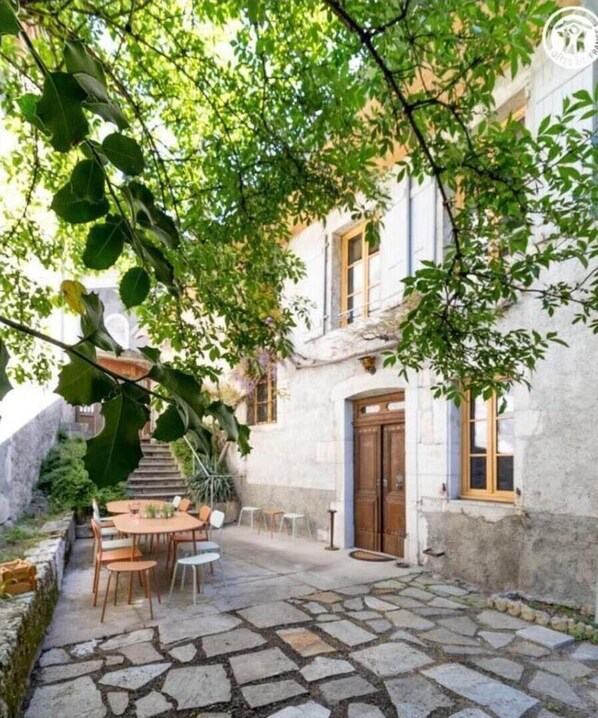 Une cour pavée de pierres, avec une table et des chaises, une porte en bois et un bâtiment à la façade en pierre.