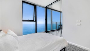 A bedroom with a large bed, a view of the sea through floor-to-ceiling windows, and a balcony with a table and chairs. - Stunning Beachfront level48 with parking /L (Surfers Paradise)