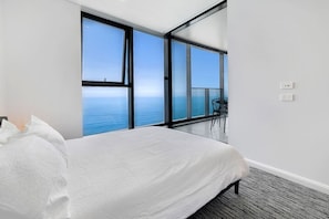 Une chambre dotée d’un grand lit, offrant une vue sur la mer grâce à de larges baies vitrées et donnant sur un balcon équipé d’une table et de chaises.