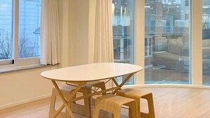 Une pièce avec une table et des tabourets en bois, de grandes fenêtres et une vue sur les bâtiments environnants.