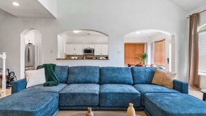 A living room with a sectional sofa, a coffee table with vases, and a kitchen in the background.