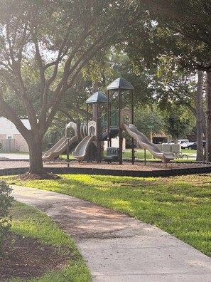 Un parque infantil con toboganes y una estructura para escalar, rodeado de árboles y un sendero pavimentado.