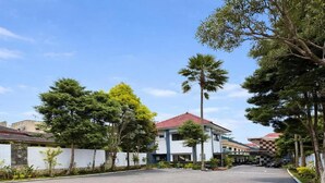 A paved walkway leading to a building with a red roof, surrounded by trees and a white wall. - Hotel Prima Asri 153 (Batu)
