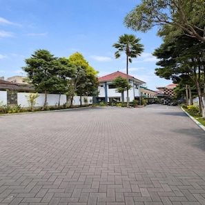 A paved walkway leading to a building with a red roof, surrounded by trees and a white wall.