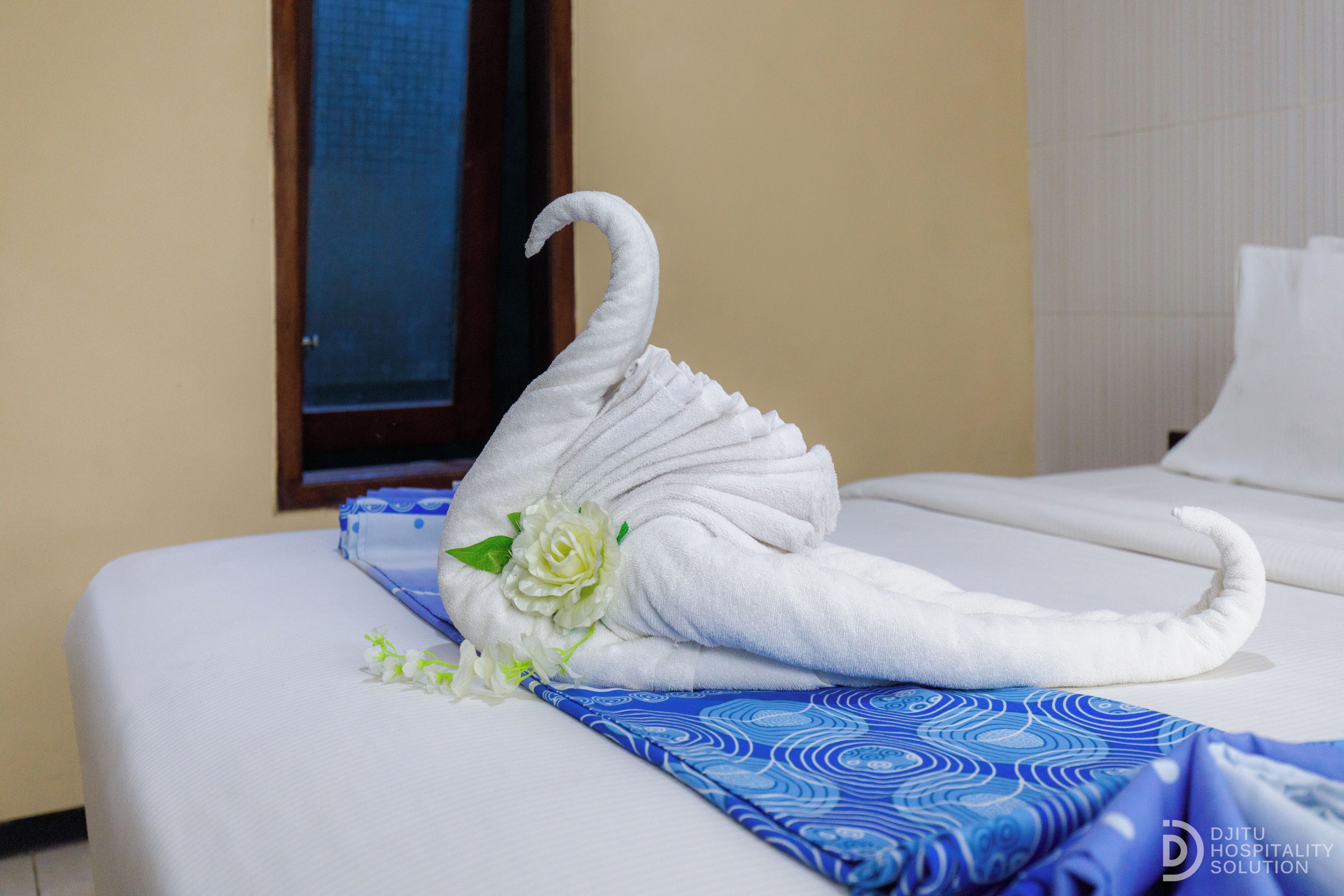 Habitación de hotel con una cama bien hecha, un centro de mesa decorativo con forma de cisne de toalla y una colcha azul estampada.