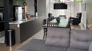 A modern kitchen with a built-in island, dining area with a table and chairs, a grey sofa, and a large window with a view of greenery.
