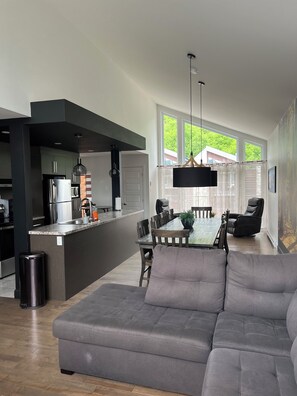Eine moderne Küche mit integrierter Kücheninsel, Essbereich mit Tisch und Stühlen, ein graues Sofa und ein großes Fenster mit Blick auf Grünflächen.
