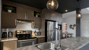 A modern kitchen with stainless steel appliances, wooden cabinets, a granite countertop, and pendant lighting.