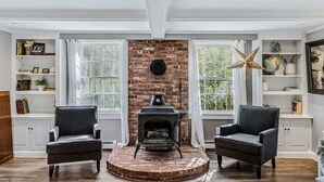 A room with a brick fireplace, two armchairs, white walls, and large windows. - Cape Cod Home in Woodstock, Pet-Friendly & Wooded (Woodstock)