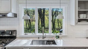 A kitchen with a double sink, stainless steel range hood, and pendant lights above a window with a forest view. - Cape Cod Home in Woodstock, Pet-Friendly & Wooded (Woodstock)