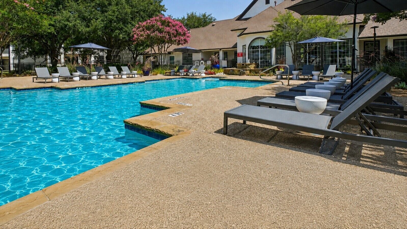 A swimming pool with lounge chairs and umbrellas, surrounded by a landscaped area with trees and a building in the background.