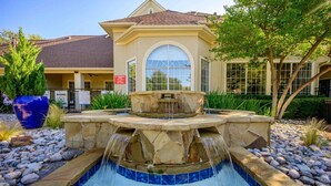 A swimming pool with a stone waterfall feature, surrounded by rocks and plants, with a building in the background.