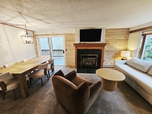 A living room with a fireplace, a TV mounted above it, a dining table with chairs, a sofa, and a small round table. - Remodeled Mountainside Condo w/Pool & Hot Tub! (Stowe)