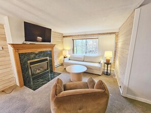 A living room with a fireplace, a sofa, a round coffee table, and a TV mounted above the fireplace. - Remodeled Mountainside Condo w/Pool & Hot Tub! (Stowe)