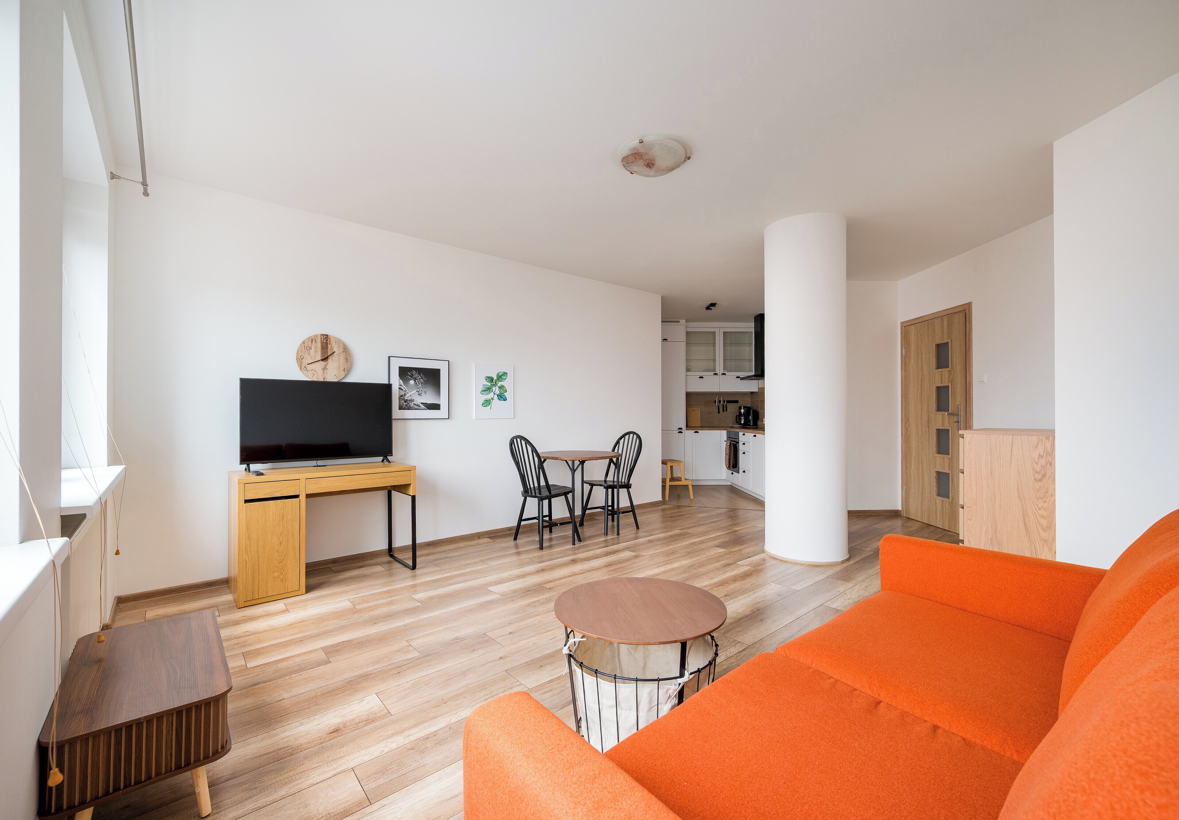 A modern living room with a sofa, a TV, a dining table with chairs, and a kitchen in the background.