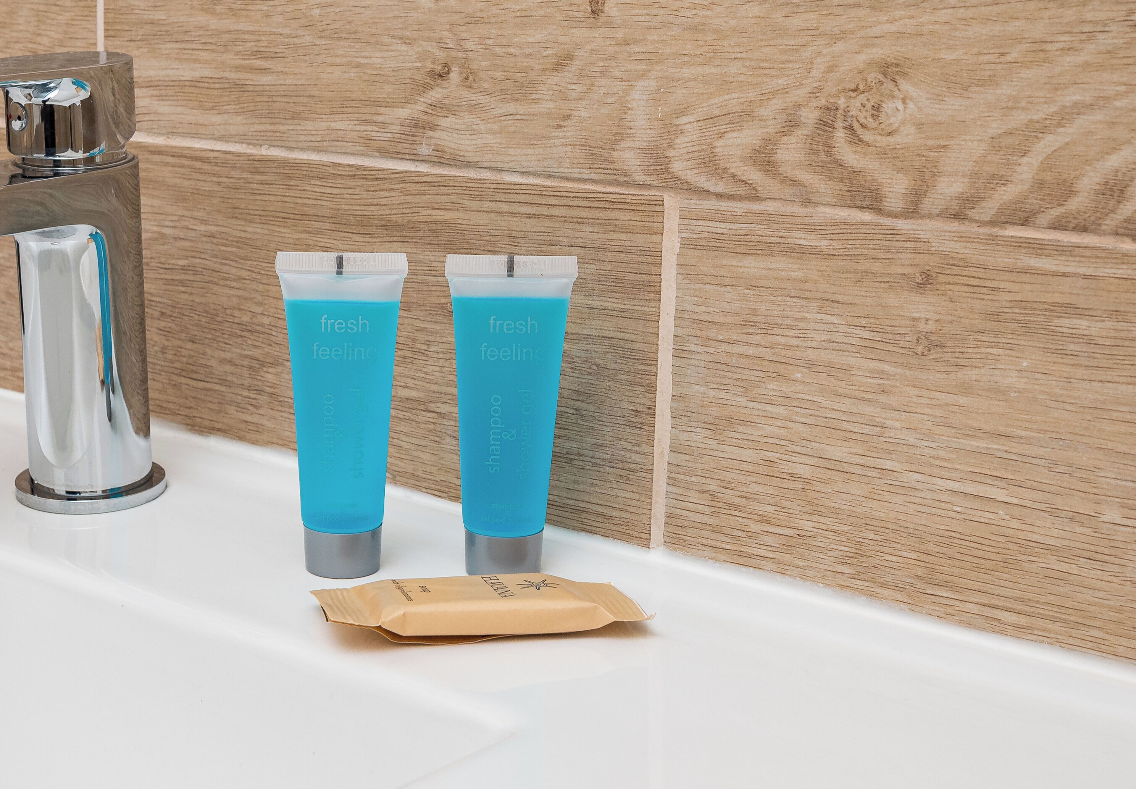 A bathroom with a modern faucet, two blue shampoo and conditioner tubes, and a brown toiletry bag on a white sink.