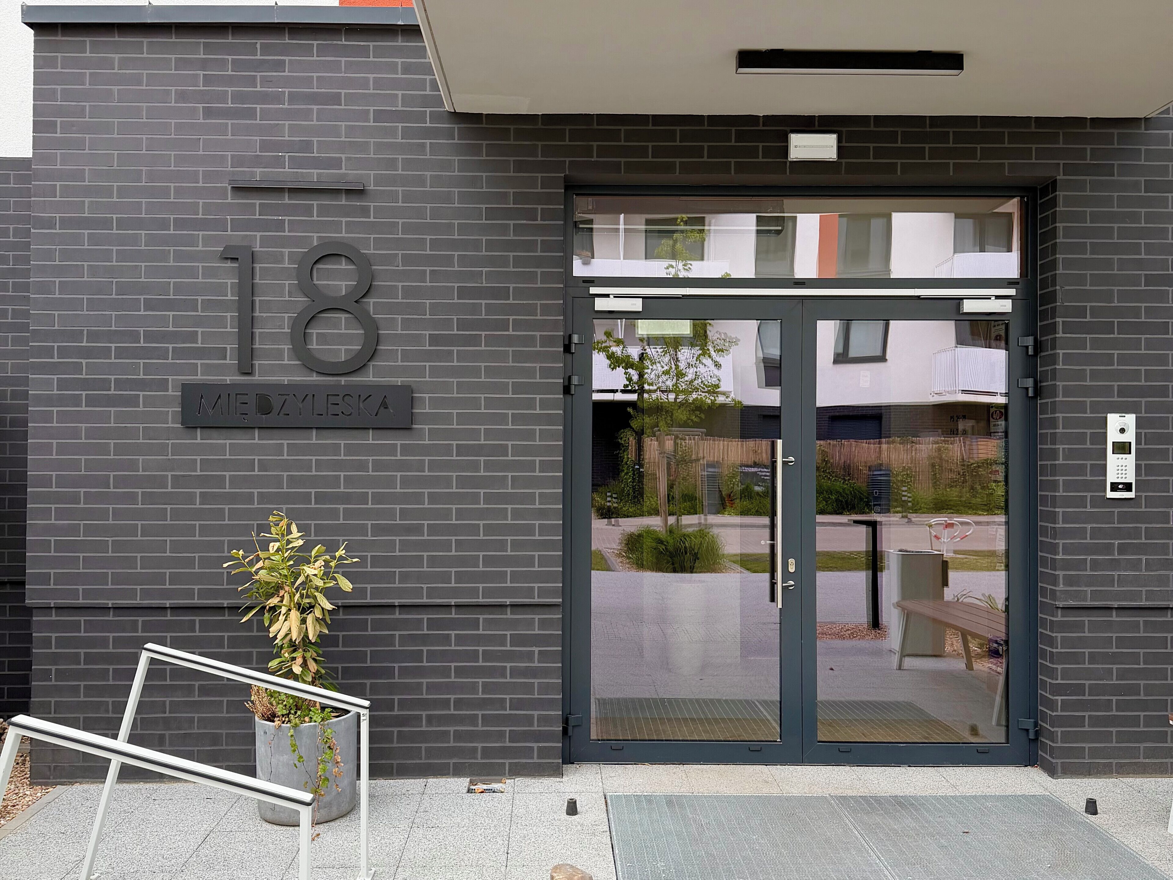 Entrada de un edificio moderno con puerta de cristal, un letrero con el número 18 y una planta en maceta.