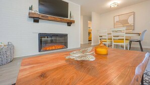 A living room with a fireplace, a flat-screen TV mounted above it, and a dining area with chairs and a table. - The Beehive New 4 Bedroom Listing Next to CNU (Newport News)