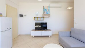 A living room with a sofa, a TV on a wooden stand, a white cabinet, a clock, and an air conditioner. - Lura Apartment 02 (Ishëm)
