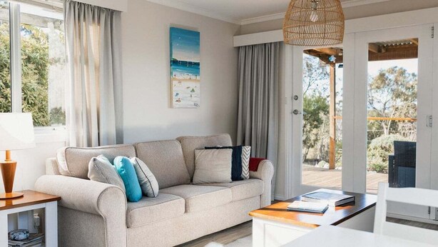A living room with a sofa, a table, a lamp, and a view of the outdoors. - Tully Cottage - a home among the gum trees (Break O'Day)