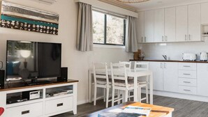 A modern kitchen with white cabinets, a dining area with white chairs, a television on a wooden stand, and a window with curtains. - Tully Cottage - a home among the gum trees (Break O'Day)