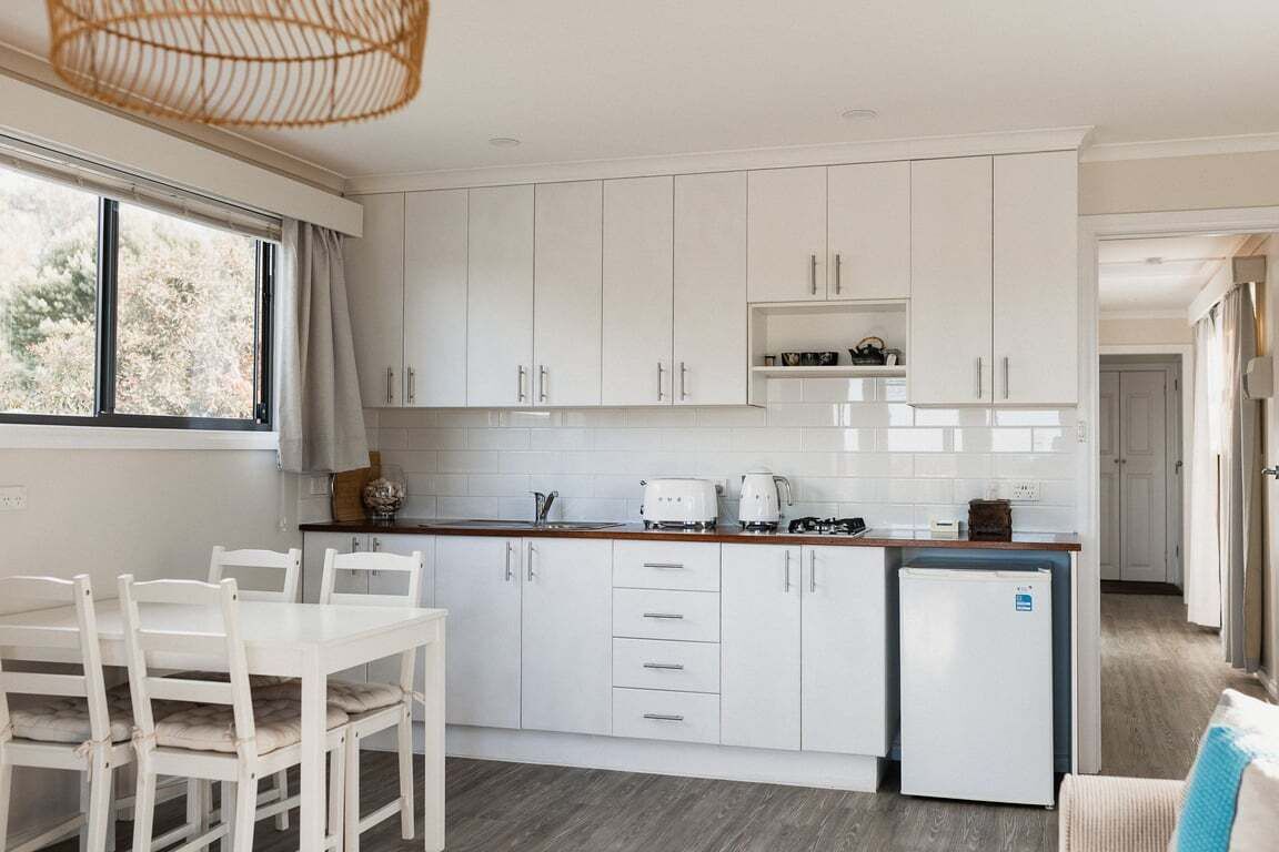 A modern kitchen with white cabinets, a dining table, and a refrigerator. - Tully Cottage - a home among the gum trees (Break O'Day)