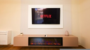 A modern living room with a mounted TV displaying 'NETFLIX,' a built-in fireplace, and a white air conditioning unit. - Sanpietro Vacation Villa 014 (Ishëm)