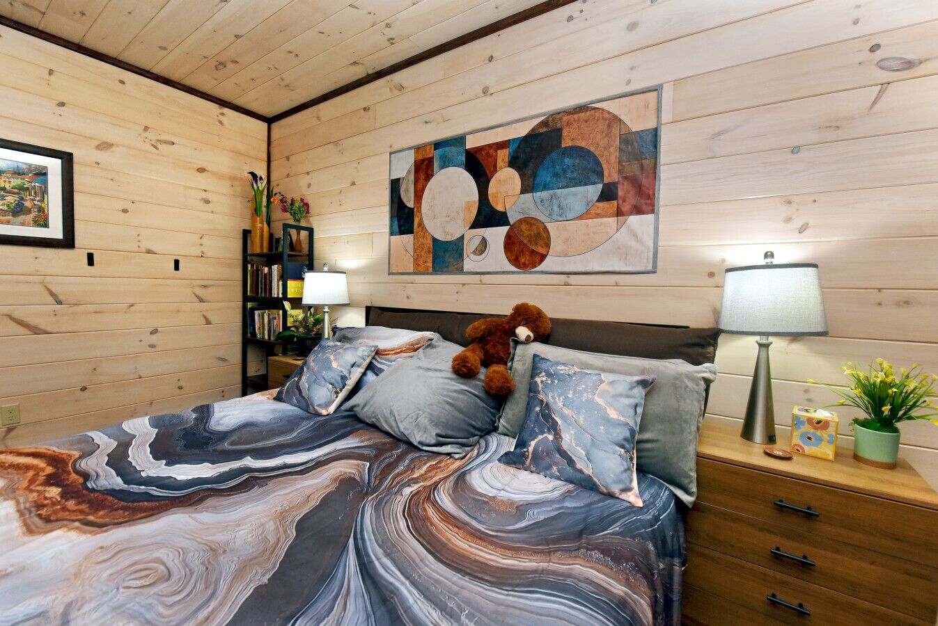 Une chambre à coucher avec un lit, des oreillers, un ours en peluche, une table de chevet, une lampe, une bibliothèque et un tableau accroché au mur.
