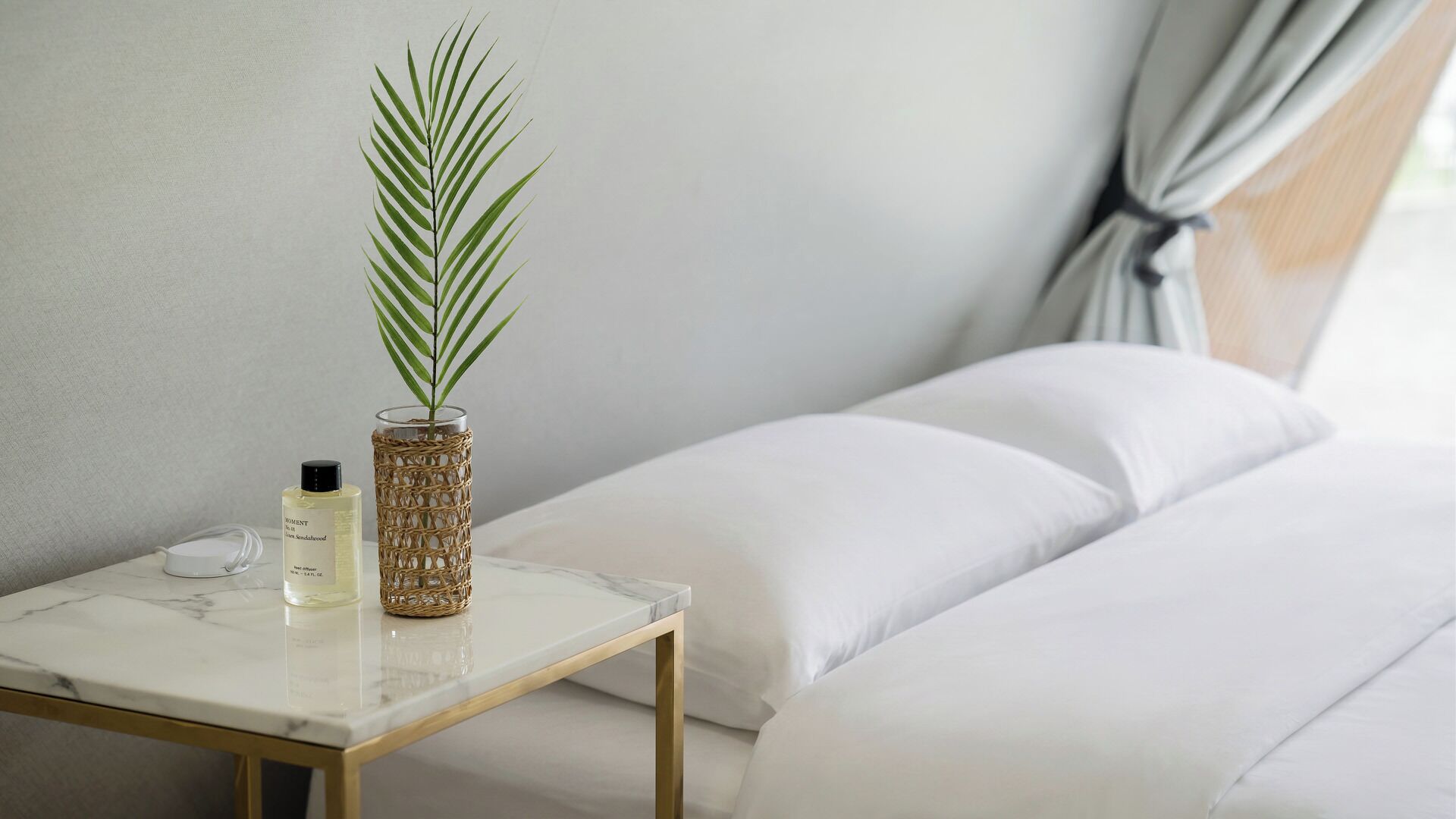 A neatly arranged bedroom with a bed, pillows, a bedside table with a bottle, a glass vase with a plant, and a curtain.