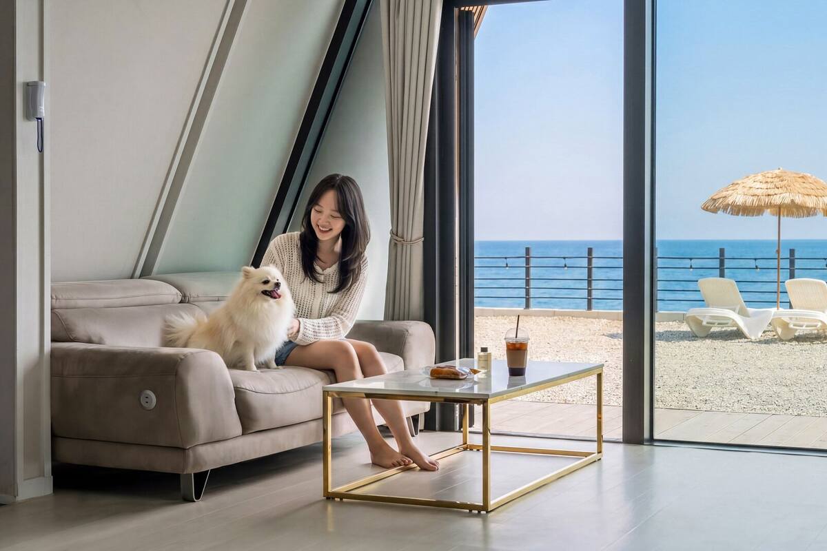 Una persona sentada en un sofá con un perro, una mesa con una bebida y vistas a la playa.