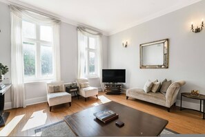 Ein modernes Wohnzimmer mit einer Couch, Sesseln, einem Couchtisch, einem Fernseher und einem großen Fenster mit Vorhängen.
