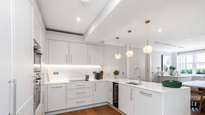 Una cucina moderna con mobili bianchi, un'isola centrale, illuminazione a sospensione e vista sull'esterno attraverso una finestra.