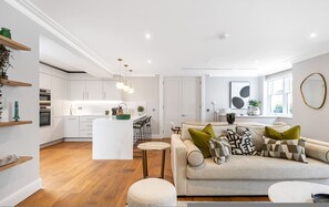 Een moderne keuken met witte kasten, een centraal eiland, hanglampen, en een zithoek met een bank en kussens.