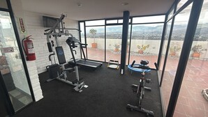 Ein Fitnessstudio mit Trainingsgeräten, einem Feuerlöscher und Blick auf eine Stadtkulisse durch große Fenster.