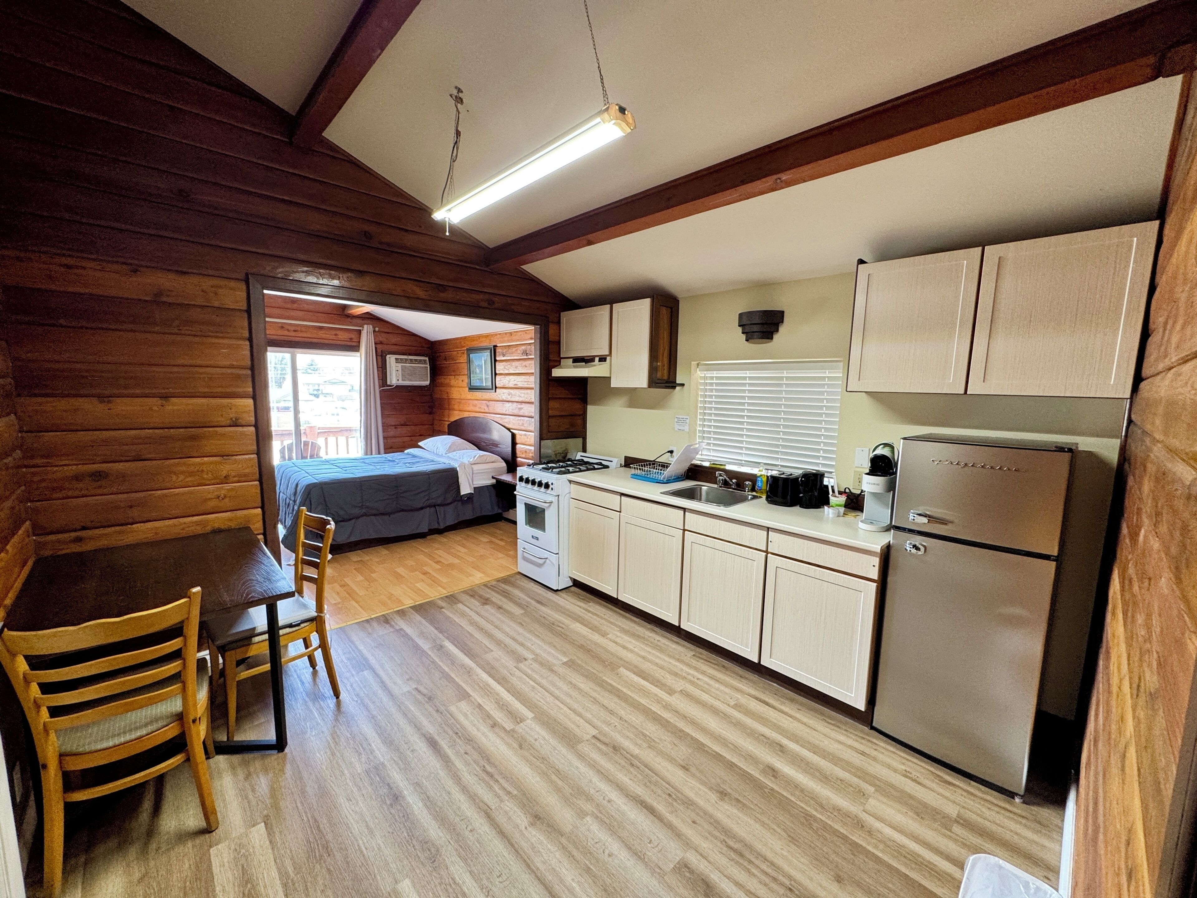 Une pièce aménagée dans le style d’un chalet en rondins, avec un lit, un plancher en bois, une cuisine équipée d’une cuisinière, d’un réfrigérateur et de meubles de rangement en bois.