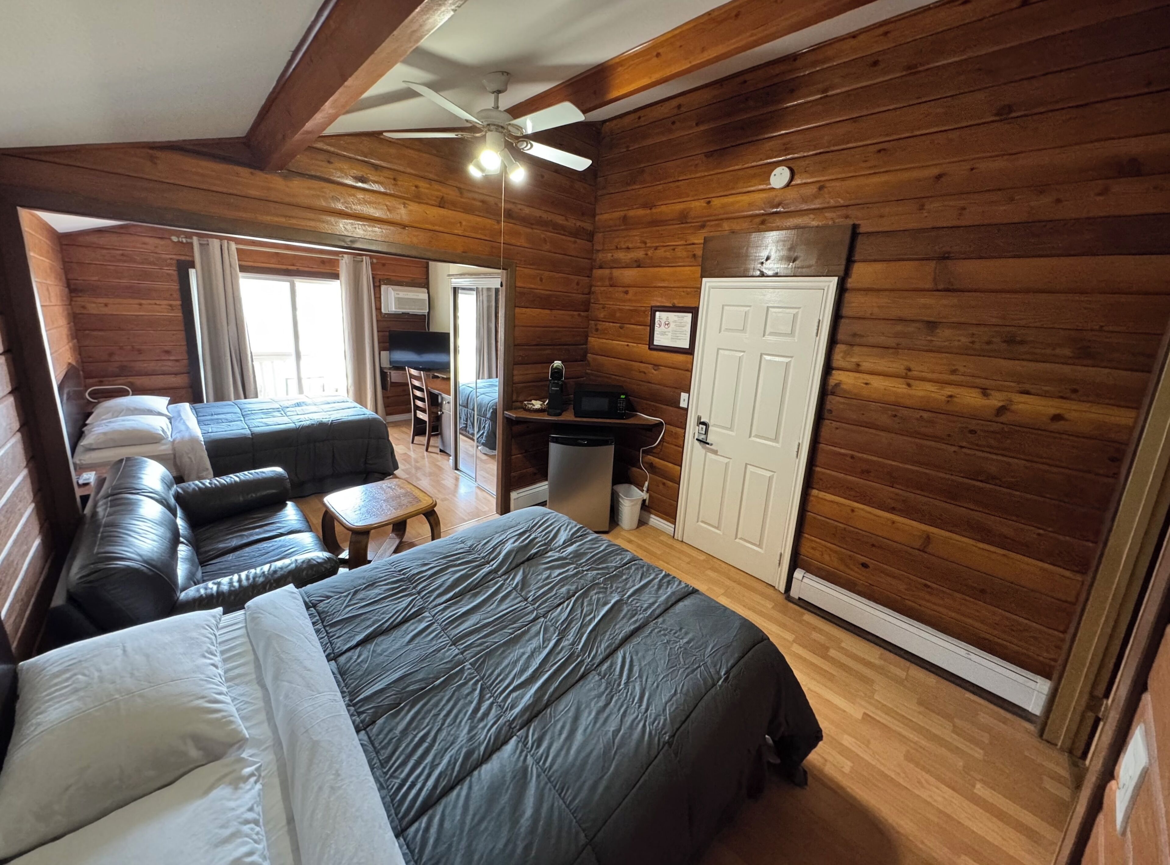 Una habitación tipo cabaña de madera con una cama, dos sofás, un escritorio con una computadora y un ventilador de techo.