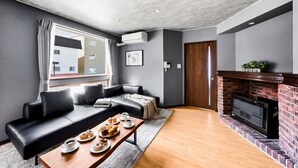 Uma sala de estar moderna com um sofá de couro preto, uma mesa de centro de madeira com doces, lareira e uma janela grande com cortinas.