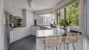 Une cuisine moderne avec des meubles blancs, un grand îlot central, des tabourets de bar et une vue sur la verdure par les fenêtres.