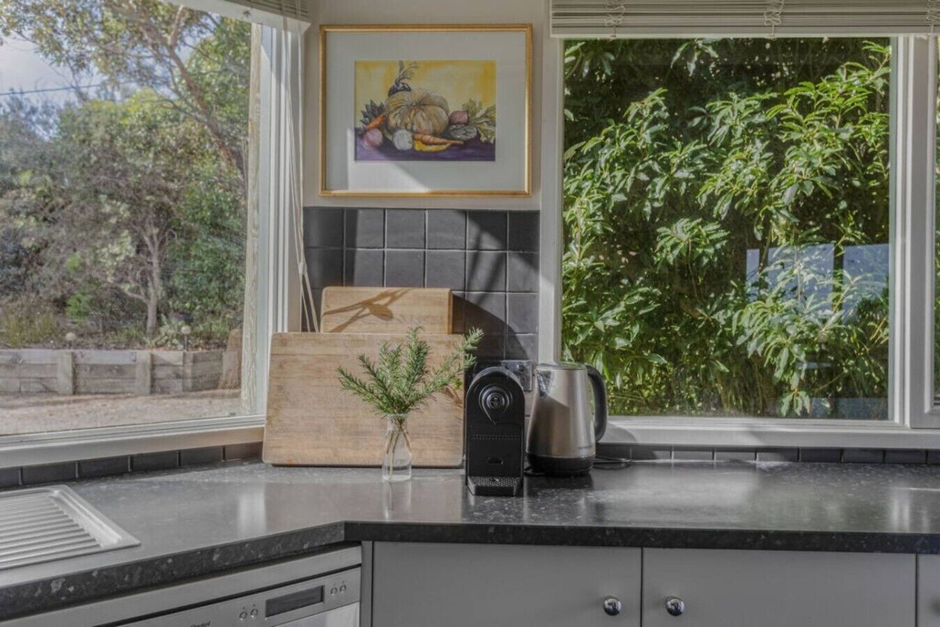A kitchen with a window, a painting, a wooden cutting board, a kettle, and a small plant.