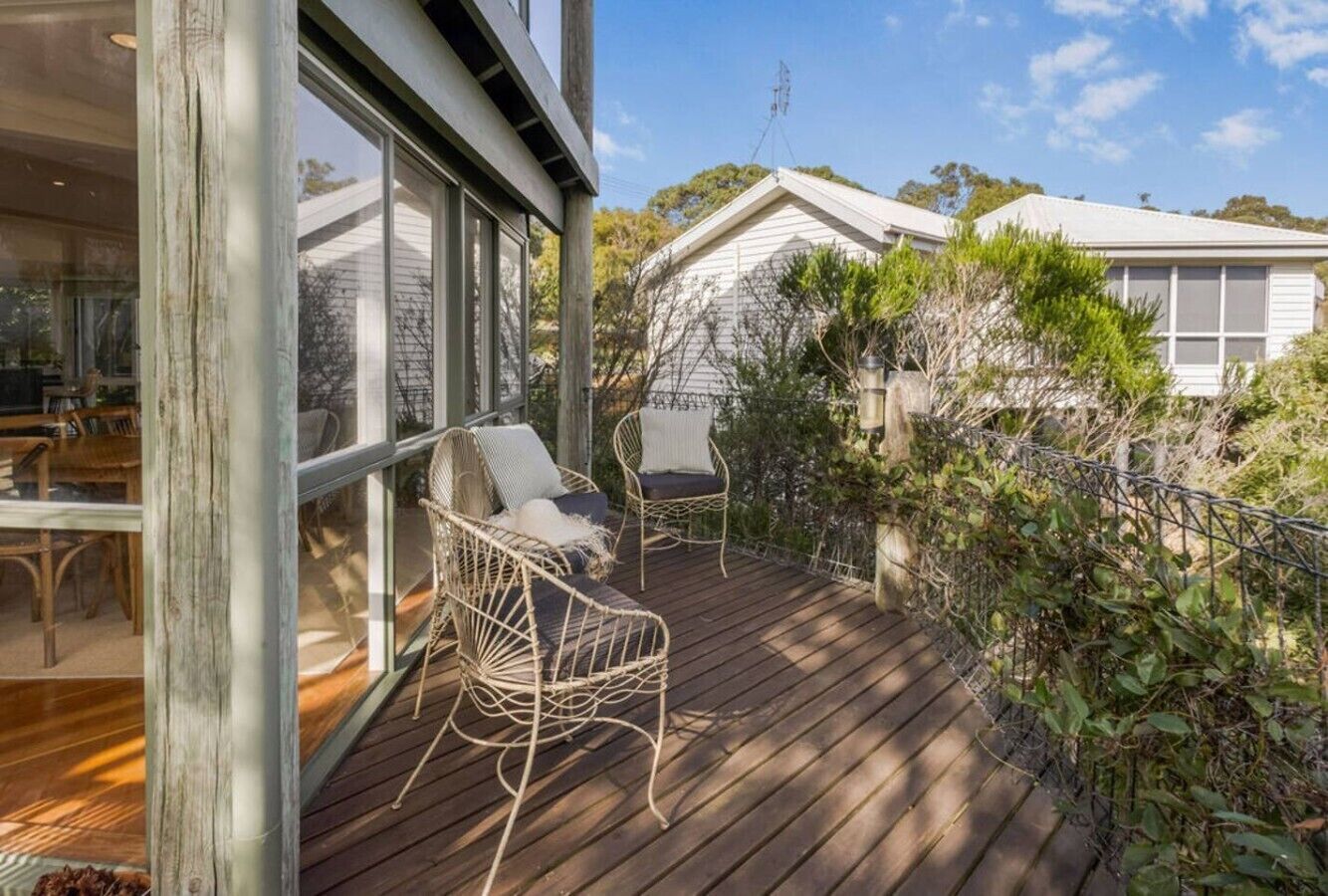 A wooden deck with wicker chairs, a glass door leading to an interior space, and a white building in the background.