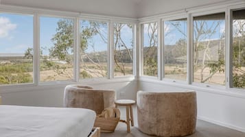 Une chambre à coucher avec un grand lit, deux fauteuils, une petite table et une vue sur un paysage naturel grâce à de grandes fenêtres.