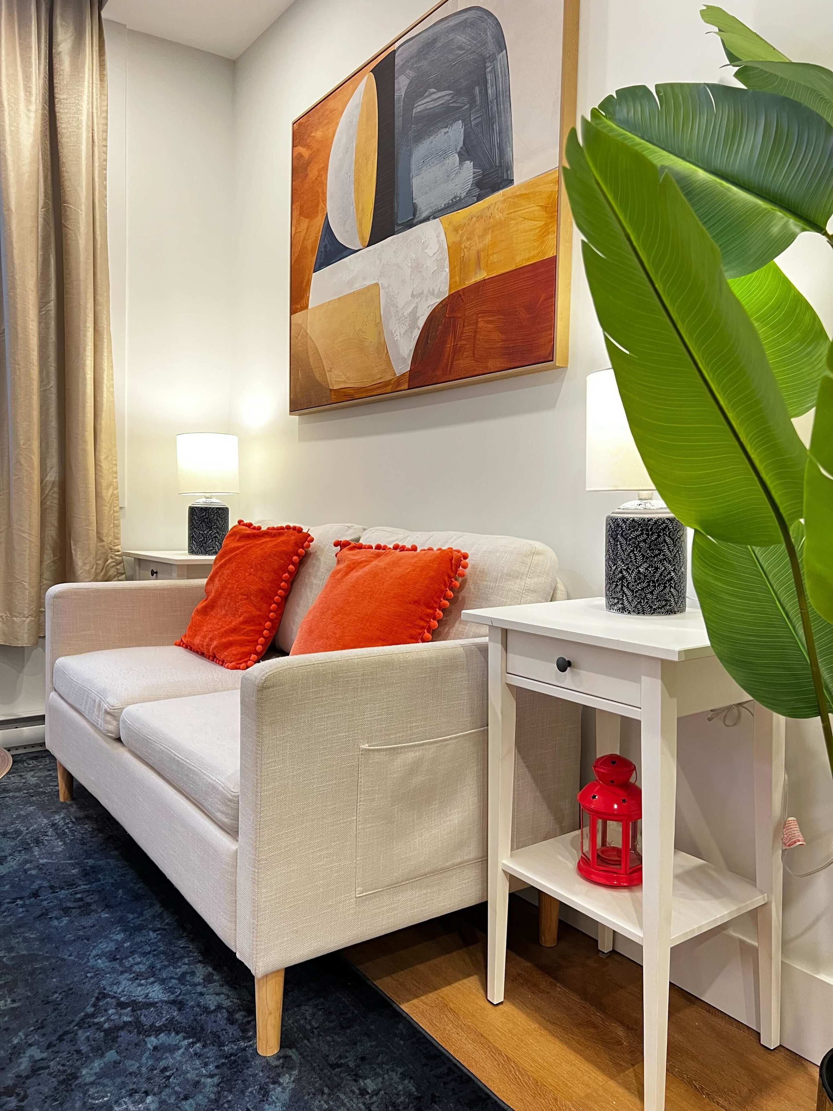 Una sala de estar con un sofá, un cuadro, una lámpara, una planta y una mesita con un farolillo rojo.