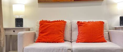 A modern living room with a sofa, two orange pillows, a glass coffee table, and a painting on the wall.