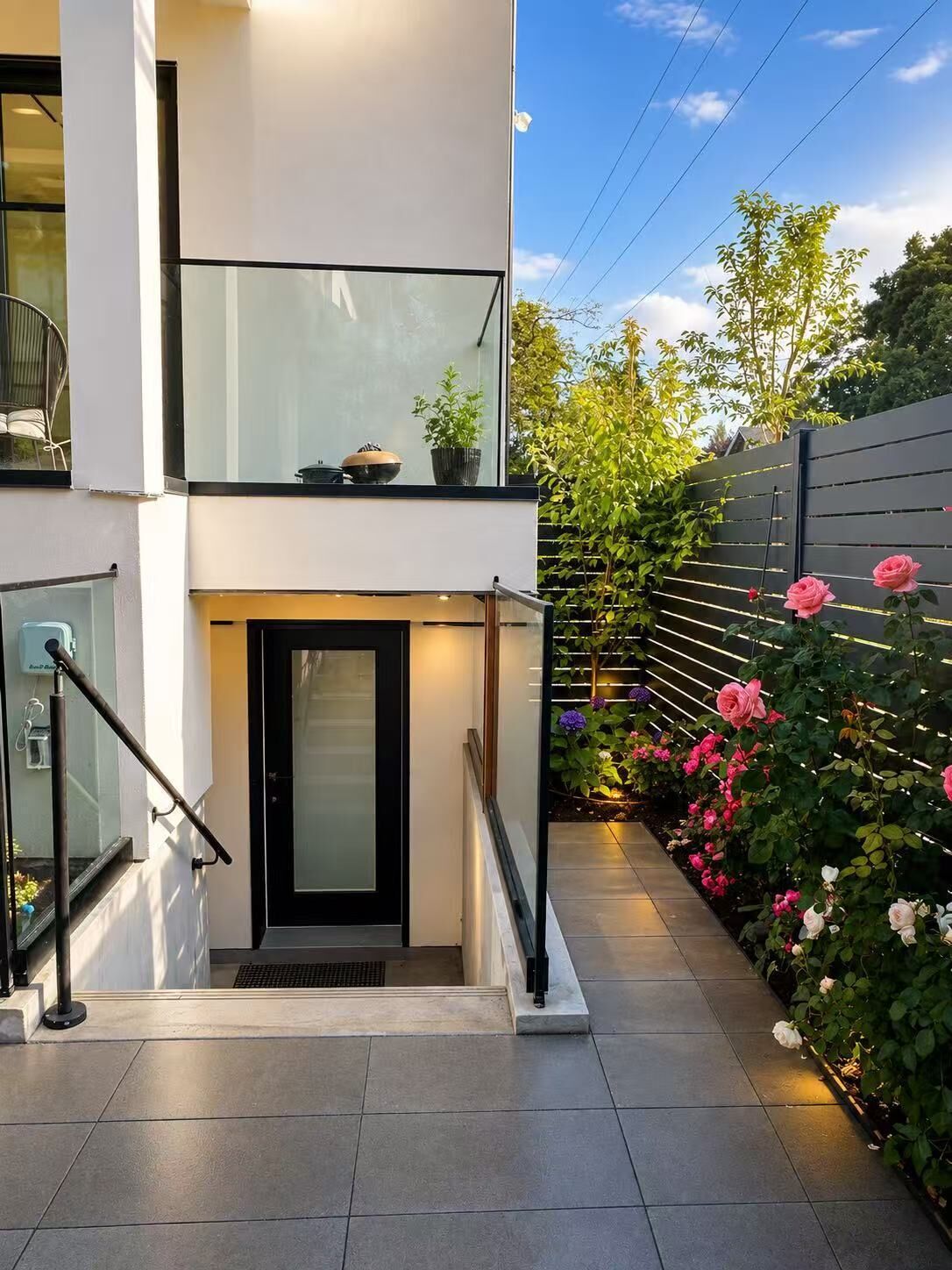 A modern two-story building with a glass balcony, a metal staircase, and a garden with roses.