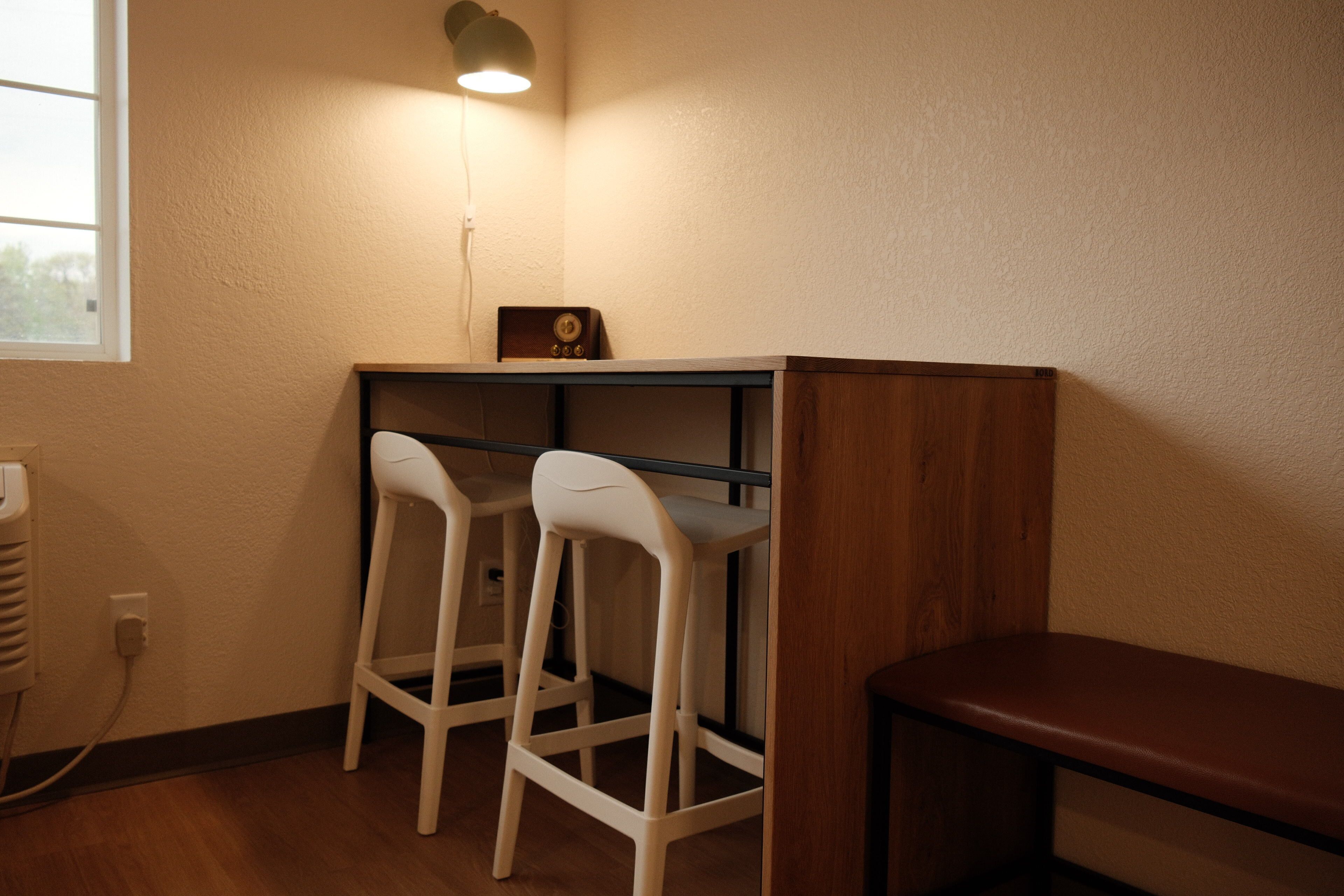 A corner room with a desk, two white bar stools, a wooden bench, a wall-mounted lamp, and a window.