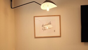 Een kleine kamer met een ronde tafel en twee stoelen, een wandlamp en een lijst met een citaat.