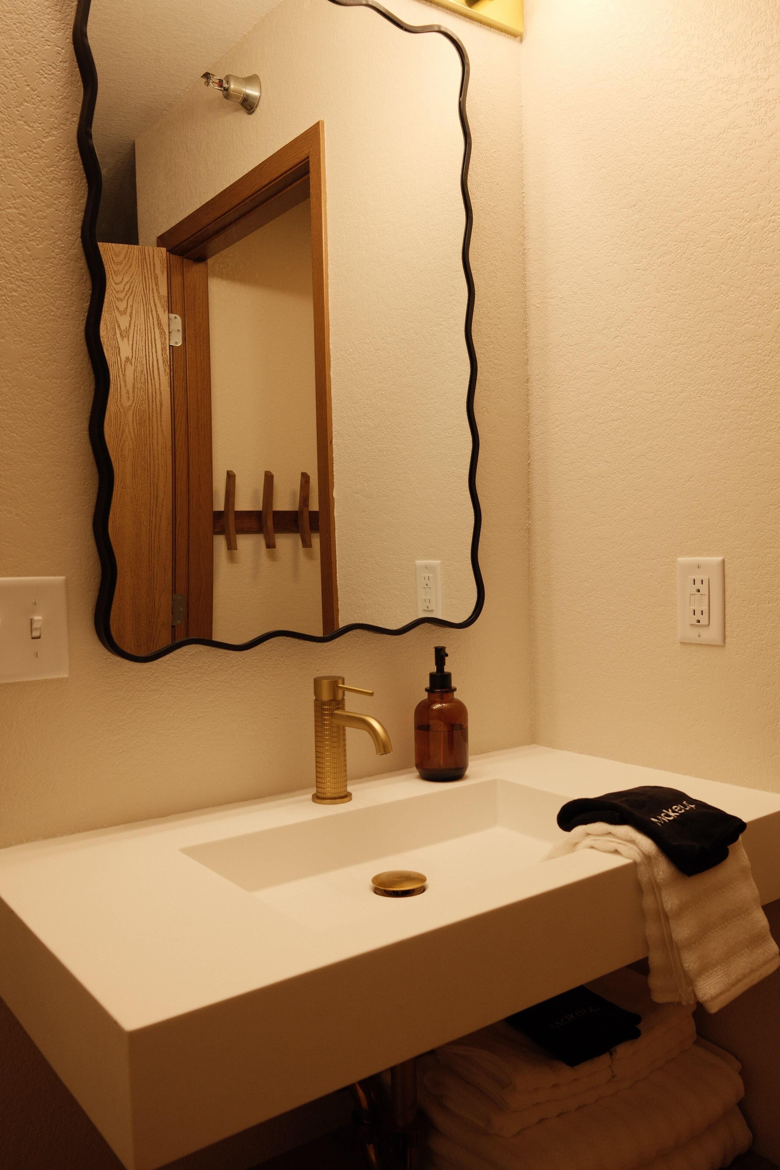 A bathroom with a white sink, a gold faucet, a mirror with a black frame, and a towel rack.