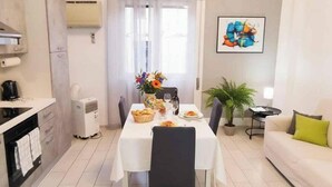 Un comedor con una mesa preparada para dos, una cocina con estufa y cafetera, una planta en maceta y un sofá con cojín.