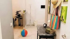 Un pasillo con suelo de madera, una mesa negra con libros y un sombrero, un bolso a rayas de colores y una lámpara de pie blanca.